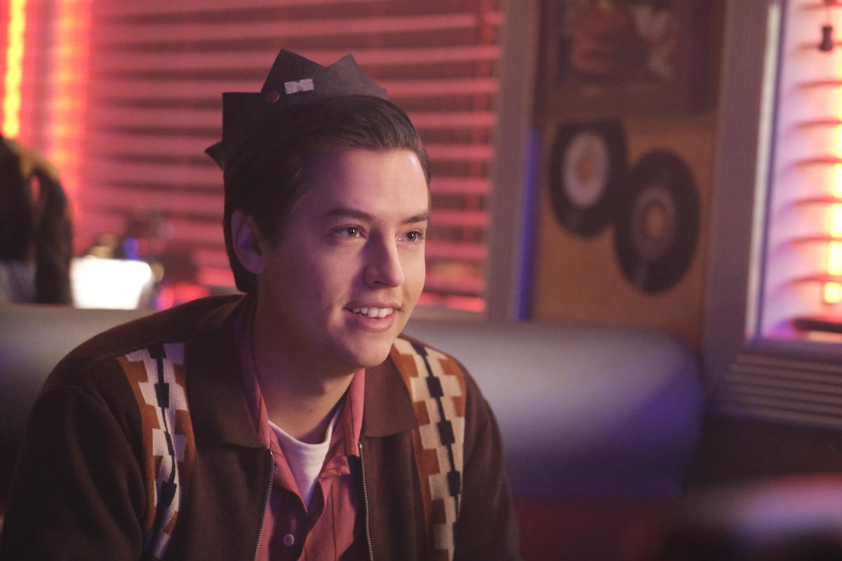 Cole Sprouse as Jughead Jones sits in a diner wearing a paper crown in Season 7 of ‘Riverdale.’