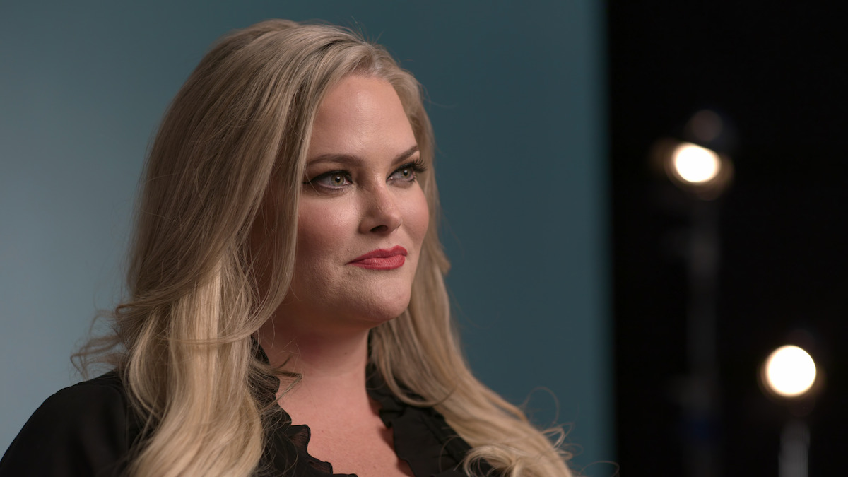 Woman with long blonde hair and black top stands in a studio with teal and black backgrounds; professional lighting visible in the background.
