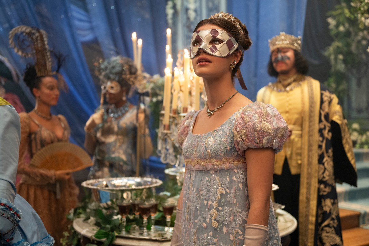 A woman in an ornate mask and elegant dress stands at a lavish, candlelit masquerade ball with elaborately dressed guests and regal decor in the background.
