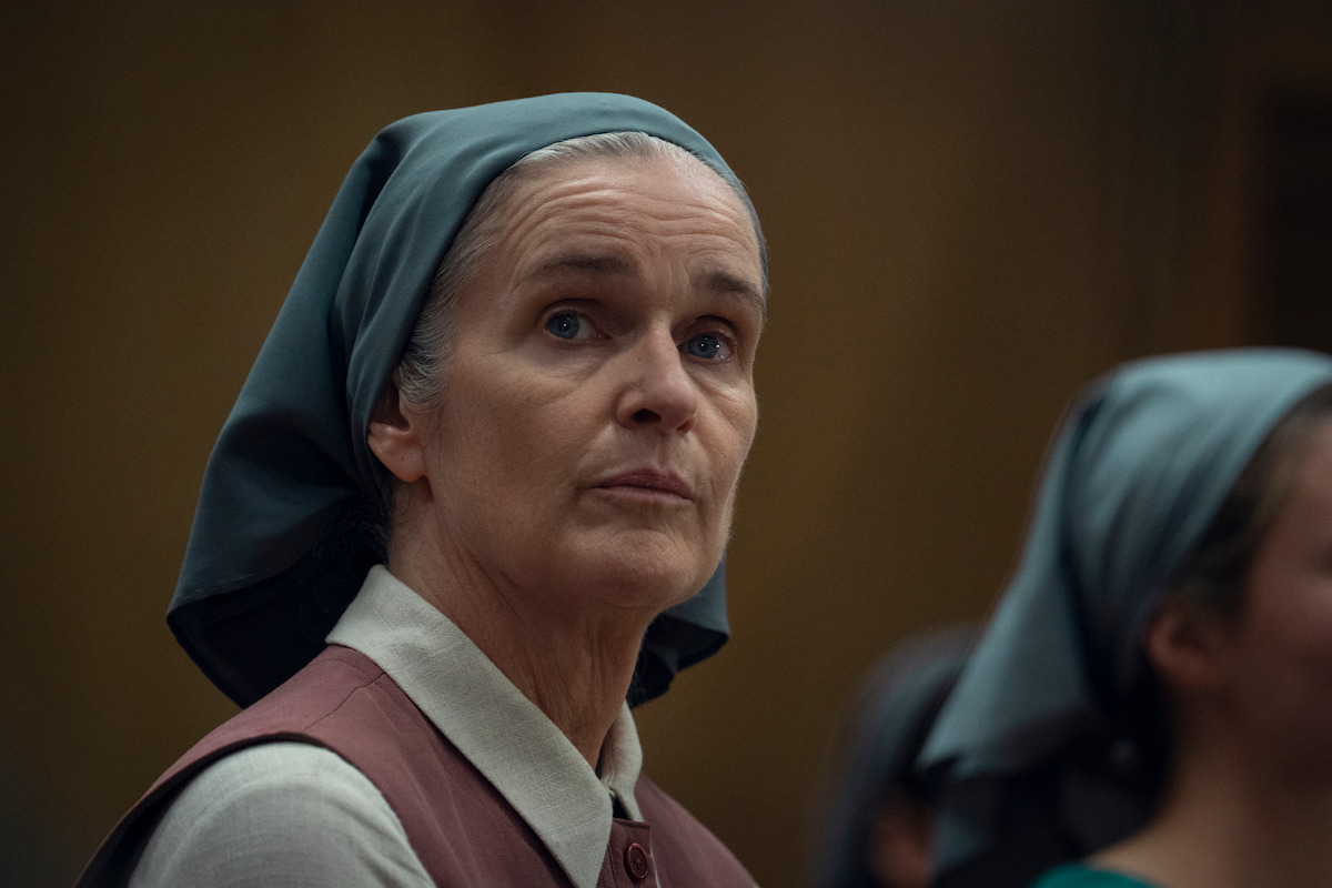 Older woman wearing a gray headscarf and modest clothing looks thoughtful in a dimly lit indoor setting, with other similarly dressed people blurred in the background.