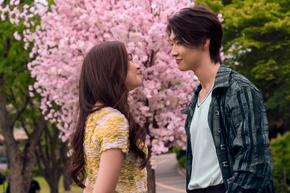 Two young adults stand facing each other and smiling beneath blooming pink cherry blossom trees in a park, surrounded by greenery on a sunny day.