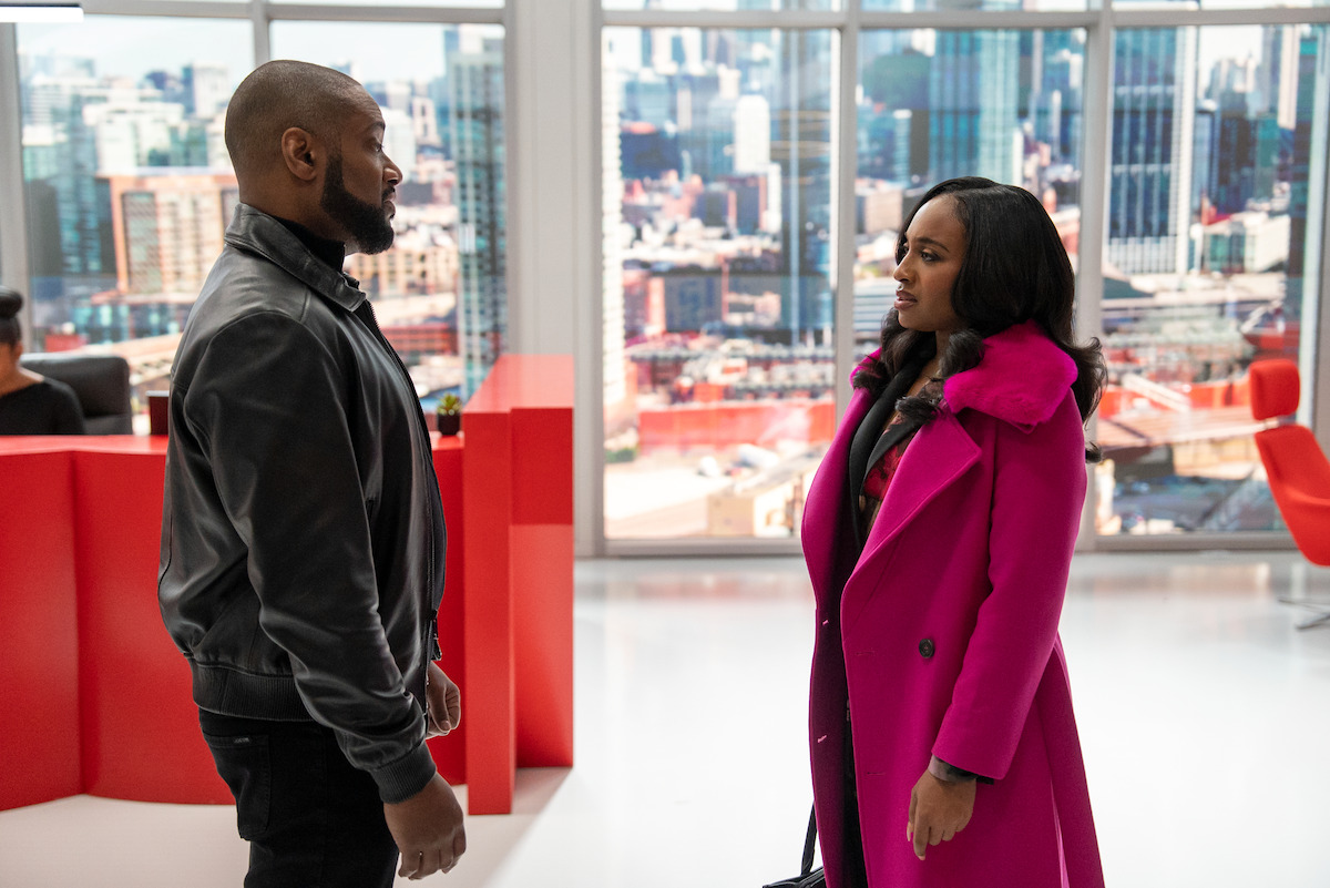 A man and woman stand facing each other in a modern, bright office space with large windows overlooking a cityscape; the mood appears serious and tense.
