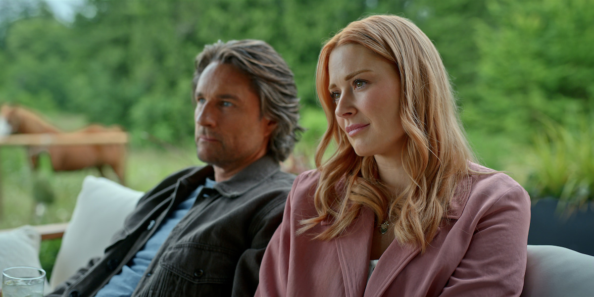A man and woman sit together on an outdoor sofa with trees and a horse in the background, appearing relaxed in a lush green natural setting.