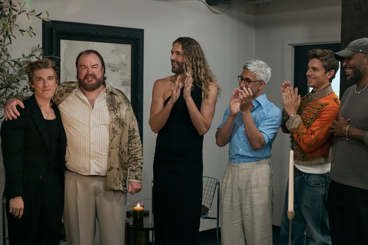 Six people standing together indoors, dressed in a mix of casual and formal clothing, smiling and interacting warmly in a well-lit modern room with art and candles in the background.