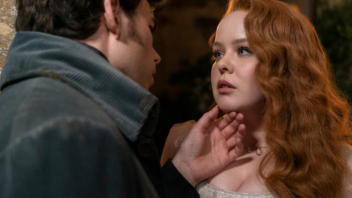 A man gently touches a red-haired woman's chin as they stand closely together indoors, both wearing period clothing. The atmosphere is intimate and emotional, with soft lighting highlighting their expressions.