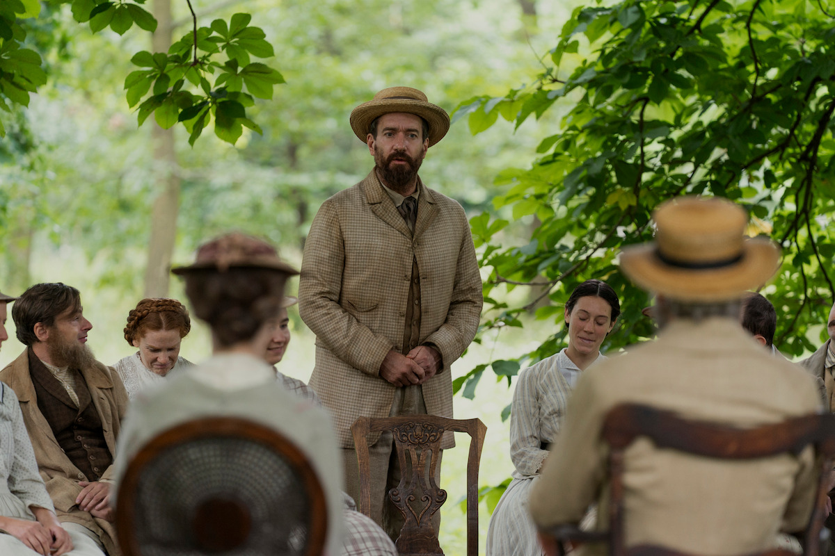 A group of people in vintage clothing sit outdoors under leafy trees, with one man standing and speaking, suggesting a historical or period gathering, calm and contemplative mood.