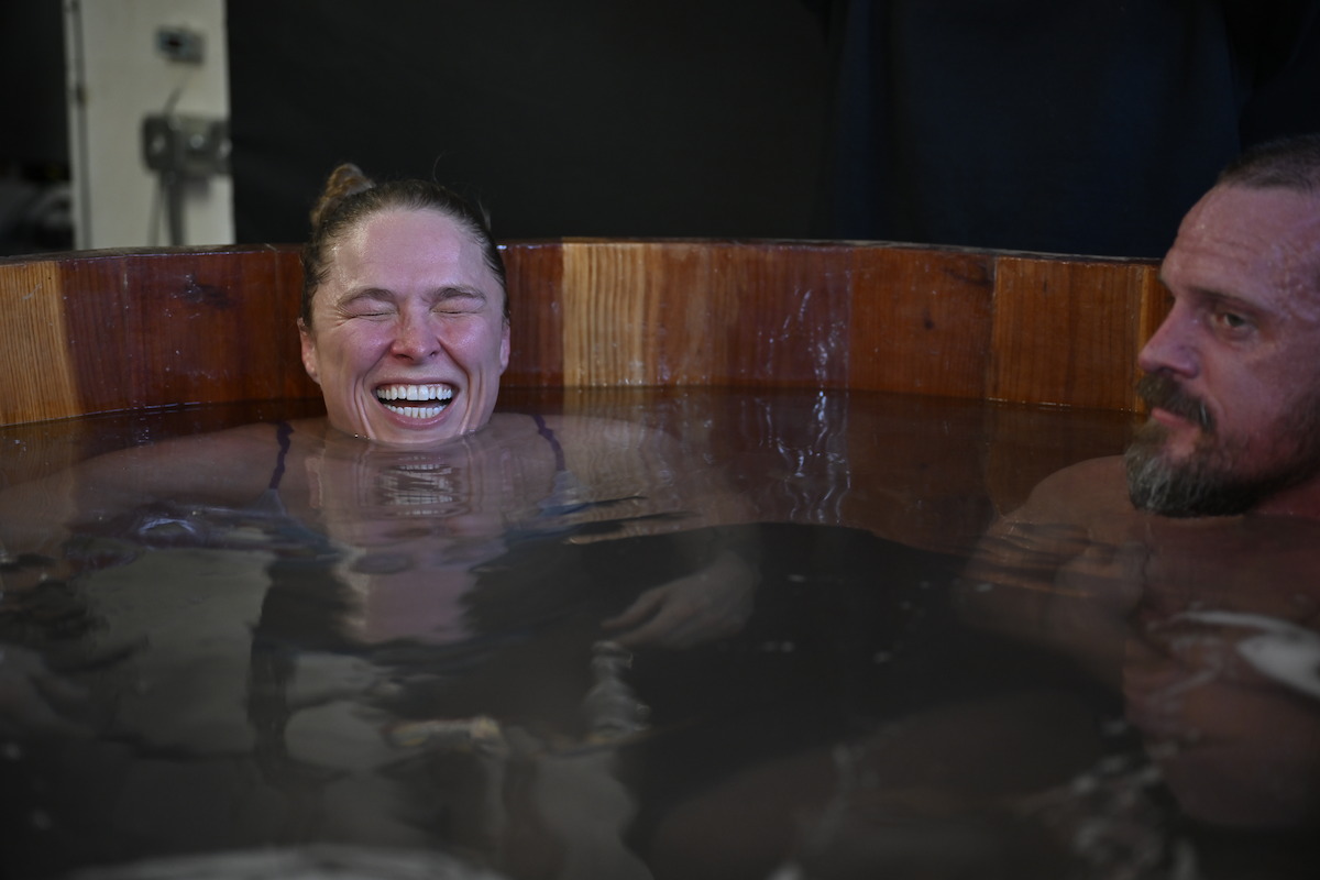 Two people sitting in a wooden ice bath, smiling and relaxed, in an indoor setting with dark background.