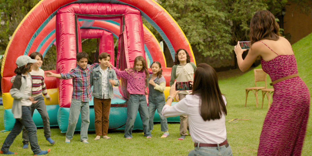 Children stand on grass in front of a bouncy house as two women take their photograph in an image from the series ‘The Accident.’