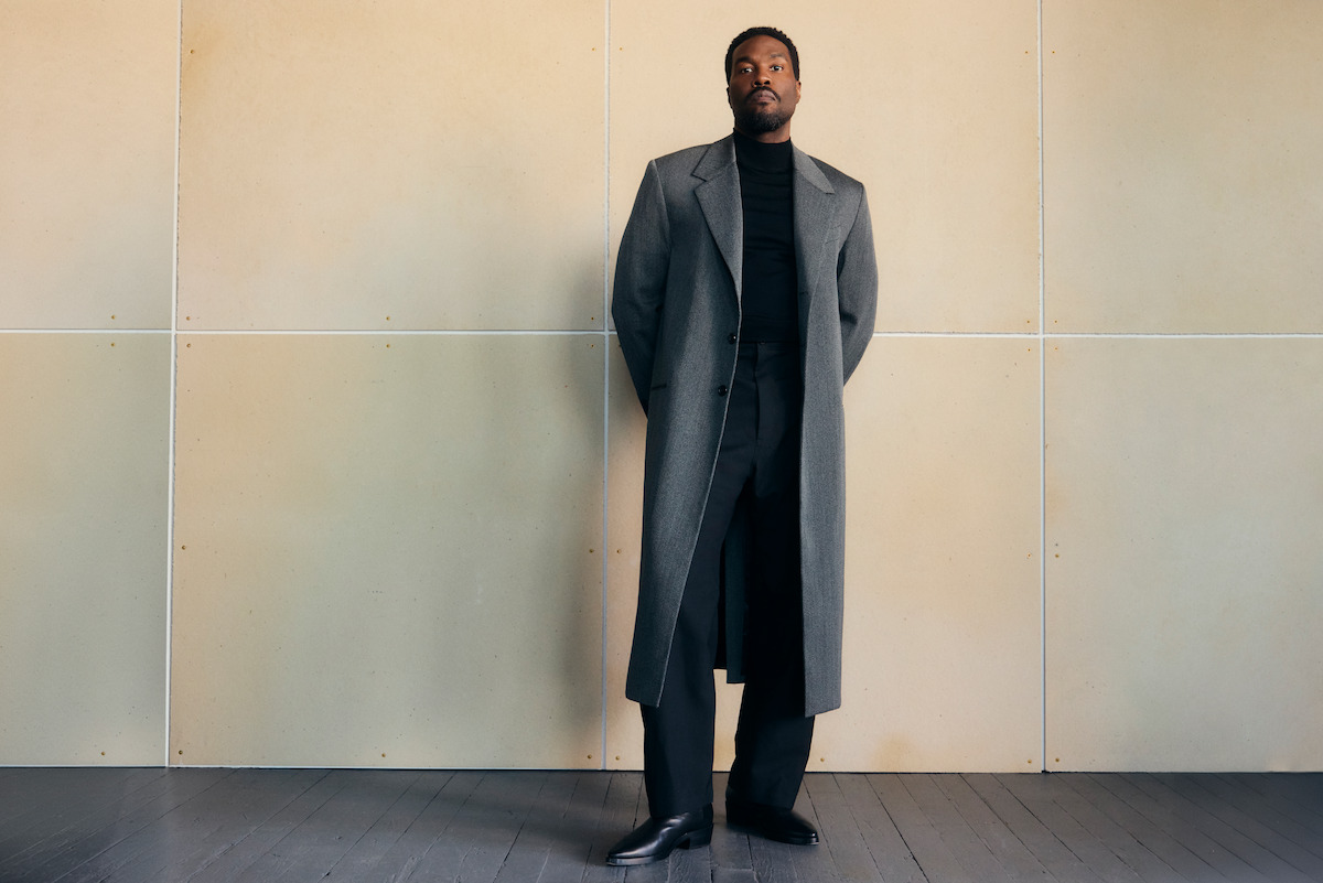 Man in a long gray coat and black turtleneck stands against a beige tiled wall in a minimalist room with gray wooden floor, hands behind his back, looking slightly upward.