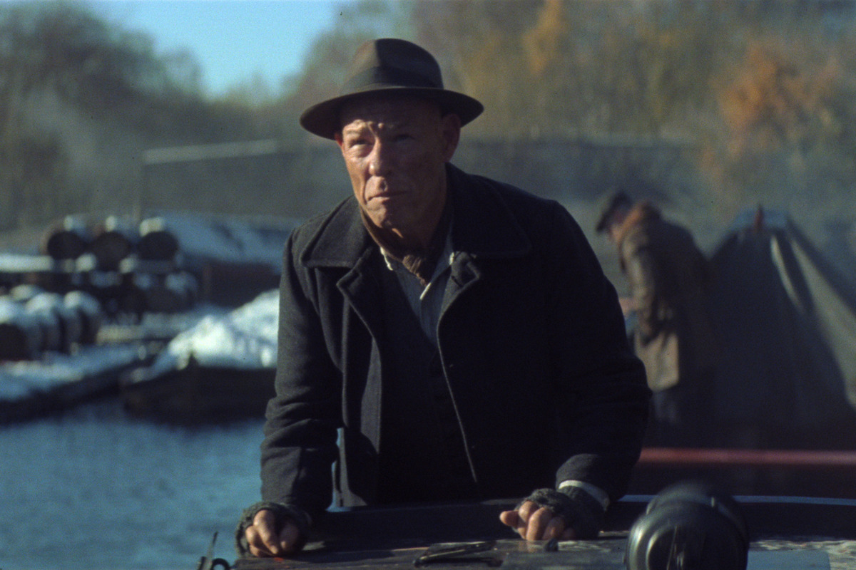 Older man in hat and coat standing on a dock or boat, with barrels and tents in the background beside a body of water on a chilly, foggy morning. Autumn trees and soft sunlight complete the outdoors rustic setting.