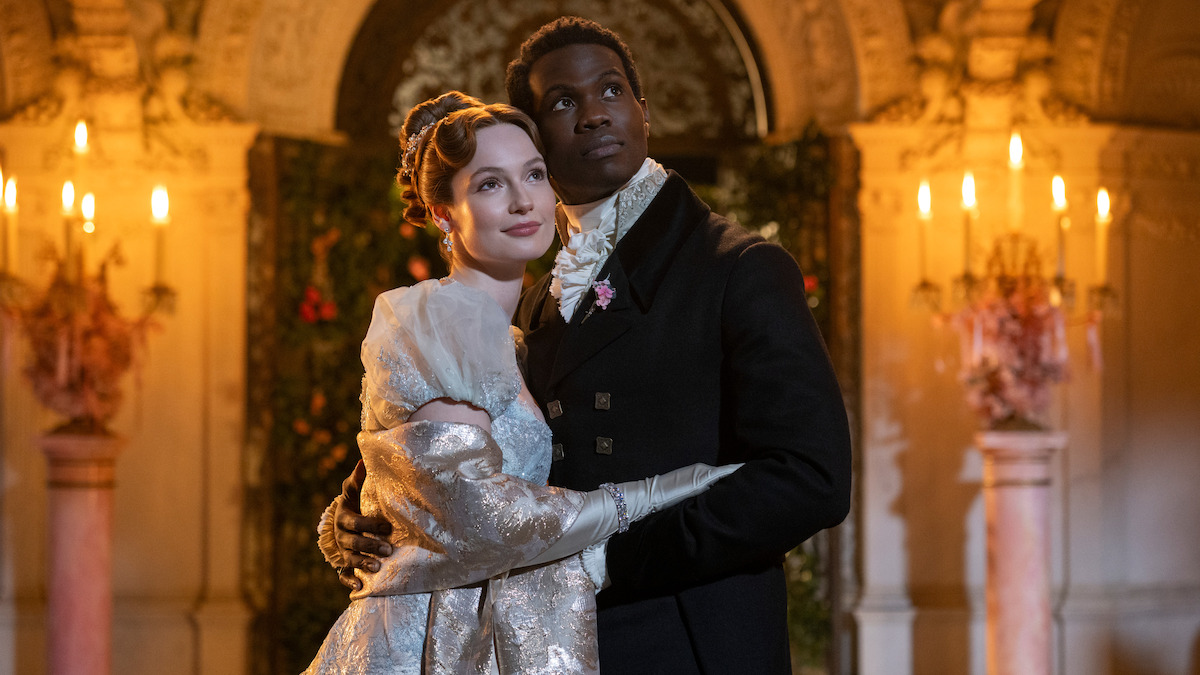 A couple dressed in elegant historical attire embrace at a lavishly decorated ballroom with glowing chandeliers and floral arrangements, creating a romantic and regal atmosphere.