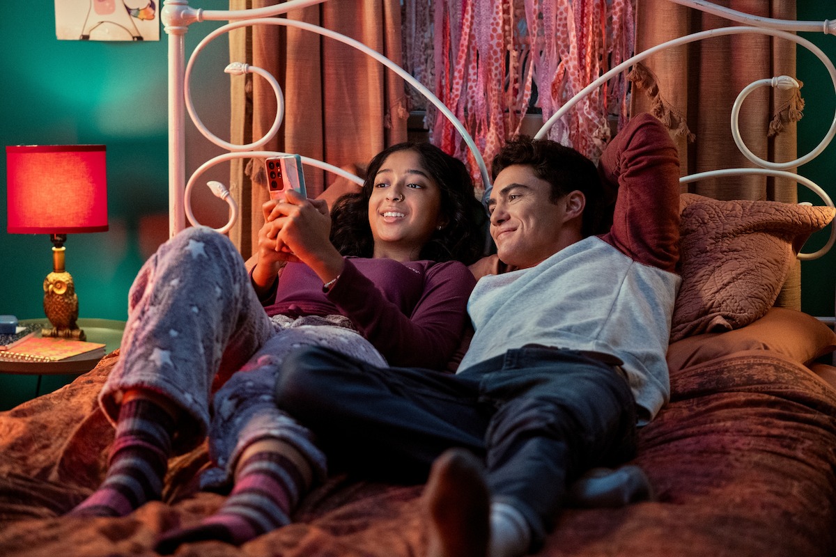 Maitreyi Ramakrishnan as Devi in Never Have I Ever Season 1 and Darren Barnet as Paxton Hall-Yoshida in Never Have I Ever Season 3 laying down on bed looking at phone.