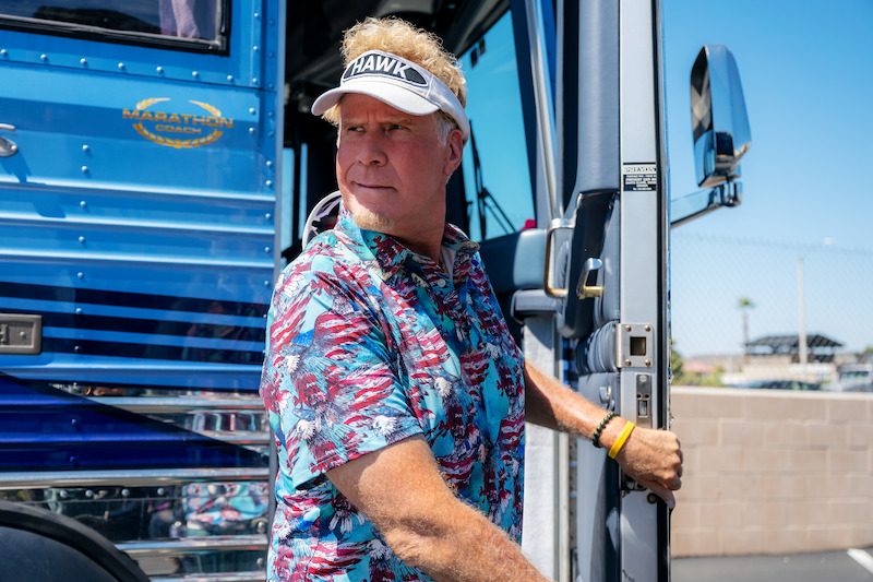 Man in a colorful shirt and visor stands by the open door of a blue bus under a clear sky, in a sunny outdoor setting with a fence and palm tree in the background.