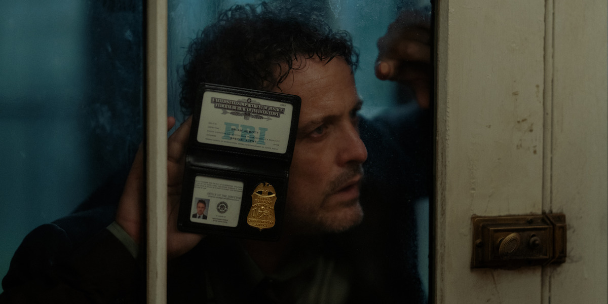 A man with curly hair shows an FBI badge and ID through a glass door on a rainy day, peering inside intently. The scene feels tense and mysterious, suggesting investigation or pursuit in a dim, somber environment.