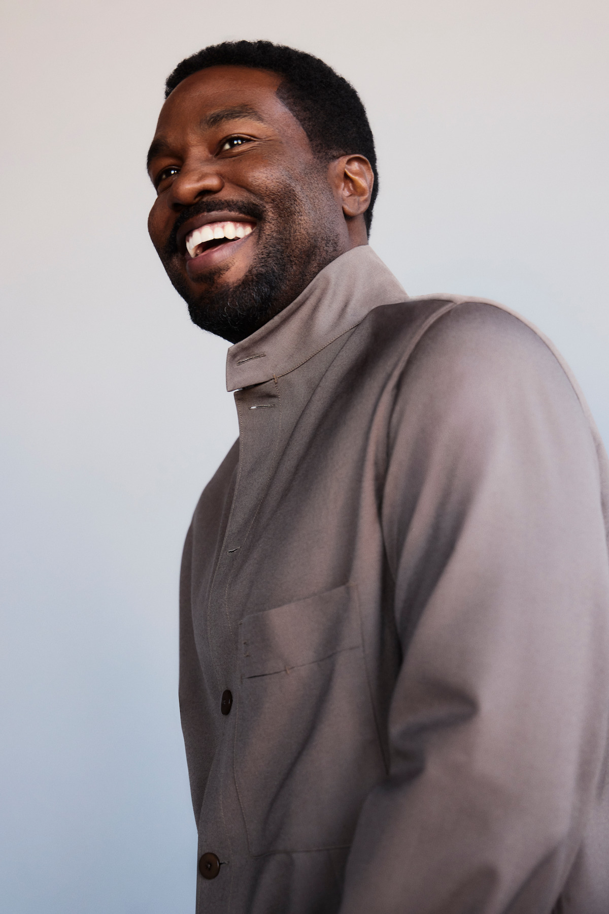 Smiling man in a gray high-collared jacket standing in front of a neutral light background, photographed from the side in a bright indoor setting.