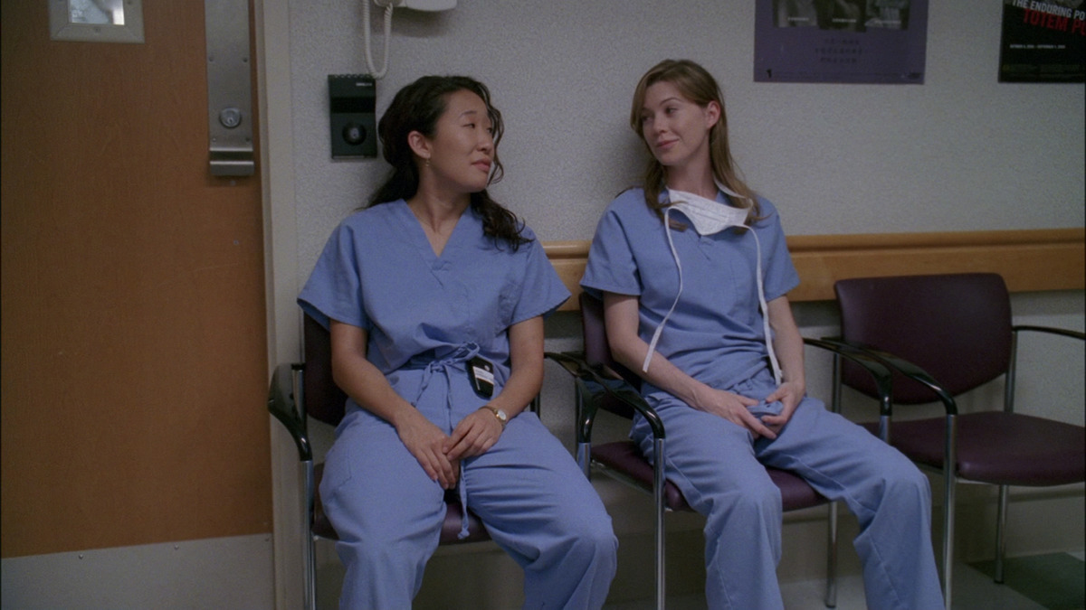 two doctors sitting next to one another in a hospital wearing scrubs. 