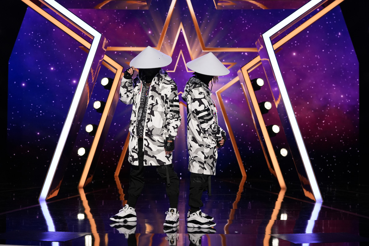 MIV in matching black-and-white outfits and conical hats stand back to back on a stage with a star-shaped purple light display, geometric lighting, and reflective floor.