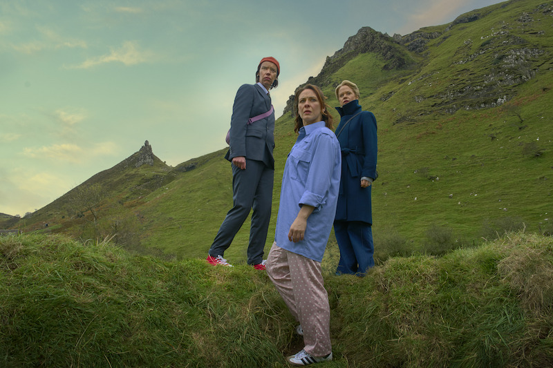 Three people stand on a grassy hillside in a rugged, mountainous landscape under a cloudy sky, dressed in casual and formal clothing, creating a contrast with the natural outdoor setting.