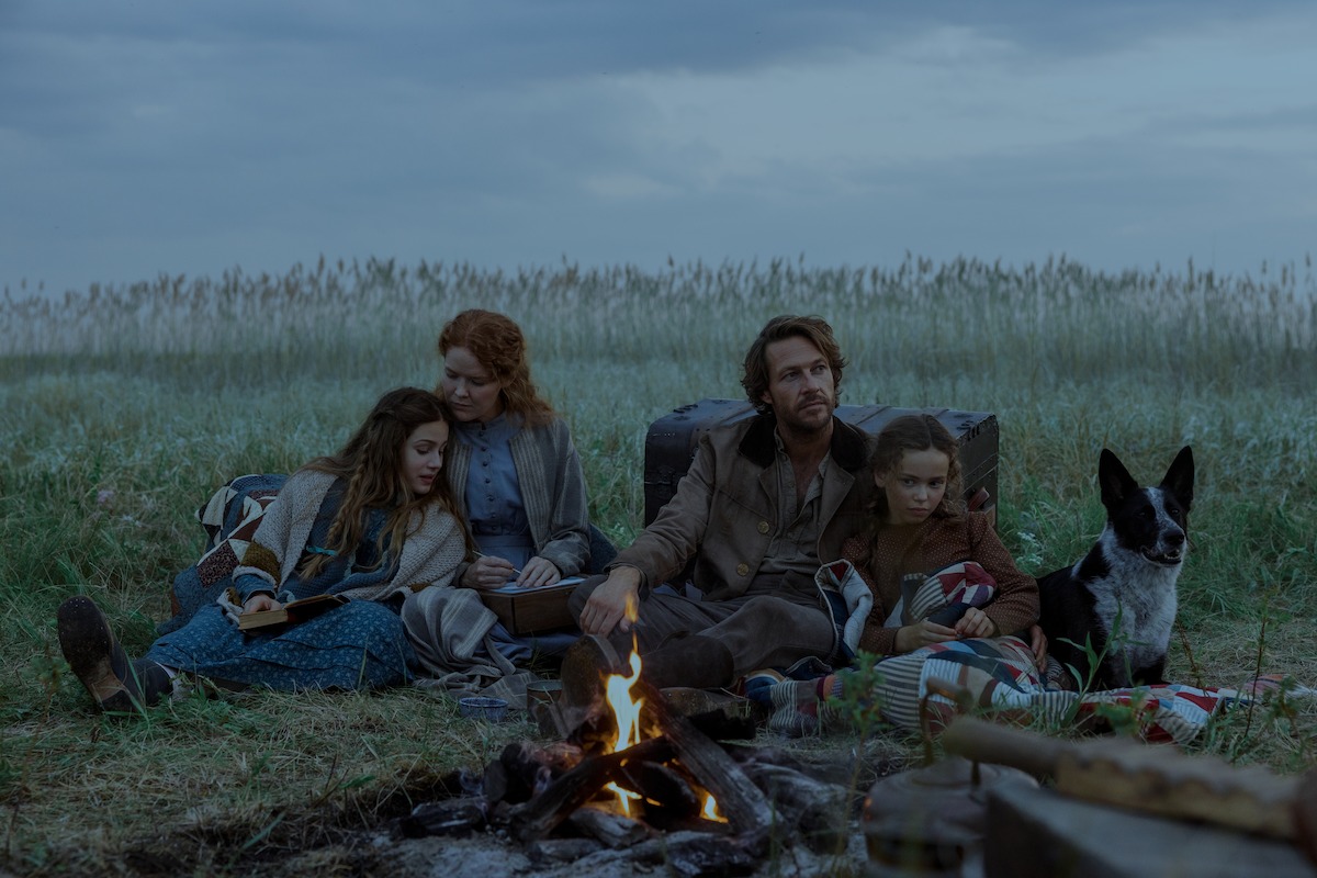 A family of four and a dog sit together by a campfire in a grassy field under a cloudy sky, surrounded by camping gear, suggesting a rustic outdoor setting and a calm, intimate atmosphere.