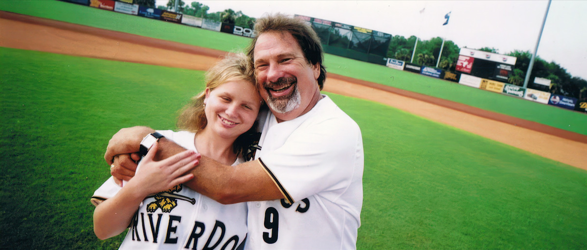 Mike Veeck hugging his daughter Rebecca Veeck on a baseball field. Both are wearing Charleston RiverDogs jerseys. 