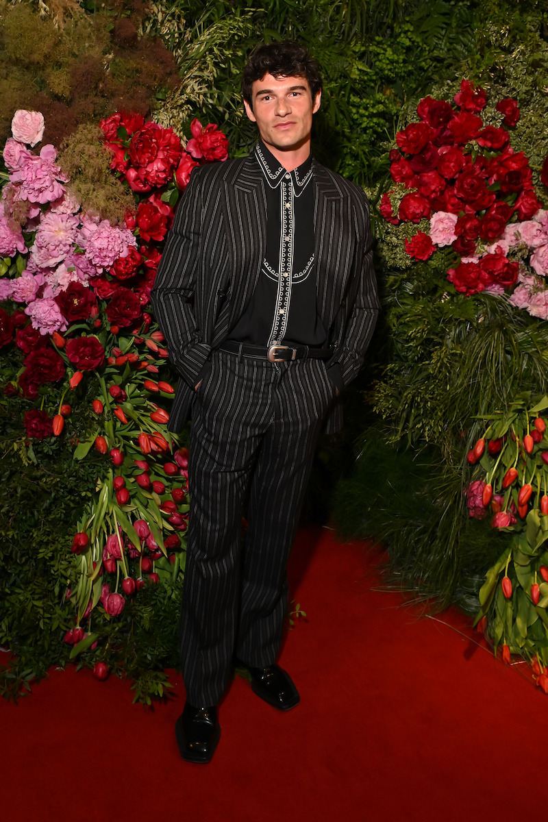 Man in a pinstripe suit stands on a red carpet surrounded by lush floral arrangements of red, pink, and purple flowers with green foliage, creating a vibrant, elegant event backdrop.