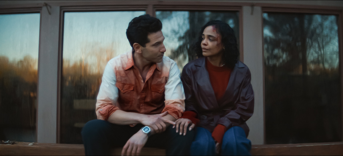 A man with blood on his shirt and woman sit closely on a wooden bench outside a building at sunset. The background shows large glass windows reflecting trees.
