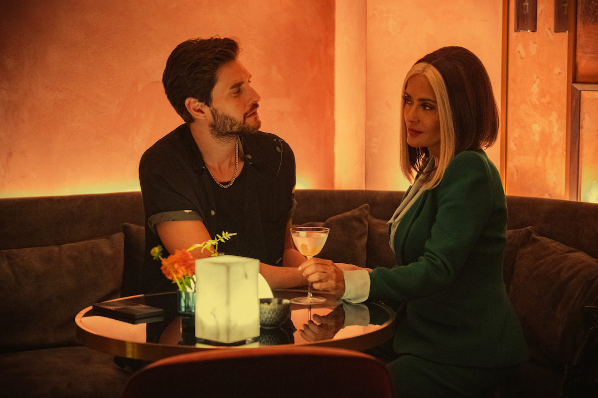 Ben Barnes as TV Mac and Salma Hayek as TV Joan sit in a corner booth at a restaurant while looking into each other’s eyes in Season 6 of Black Mirror.