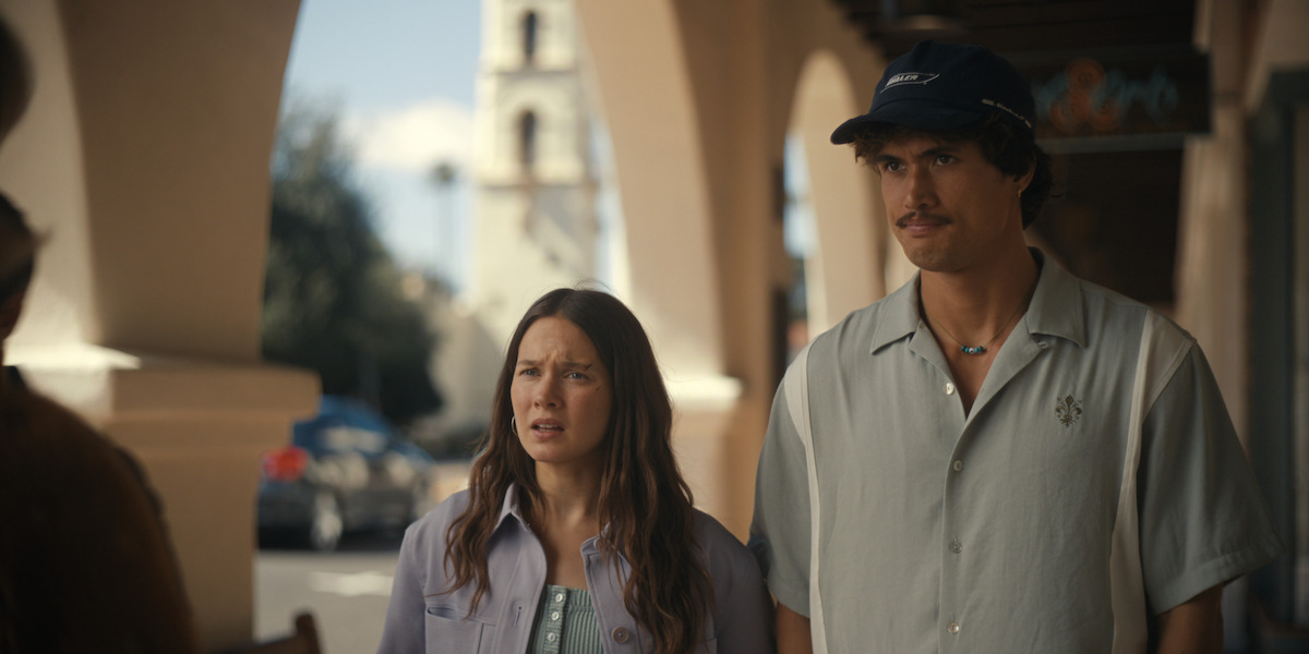 A woman and a man stand under arched walkways outside, looking concerned. The background features cars, trees, and a tower, suggesting a city or town setting on a sunny day.