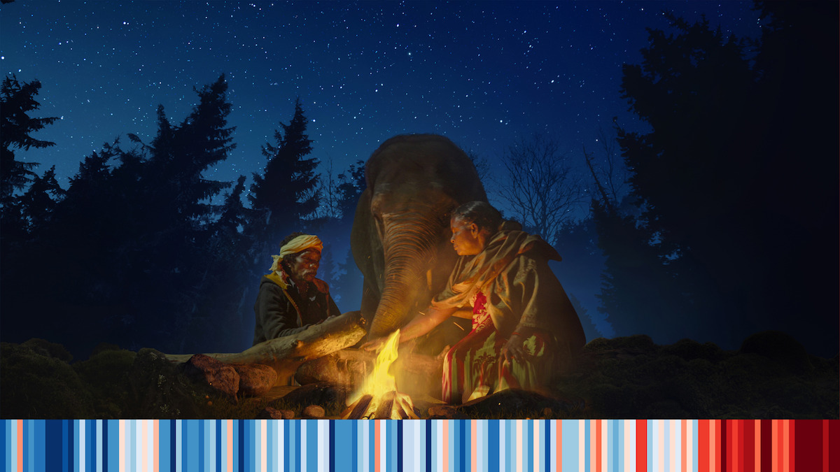 Two people and an elephant sitting around a campfire. 