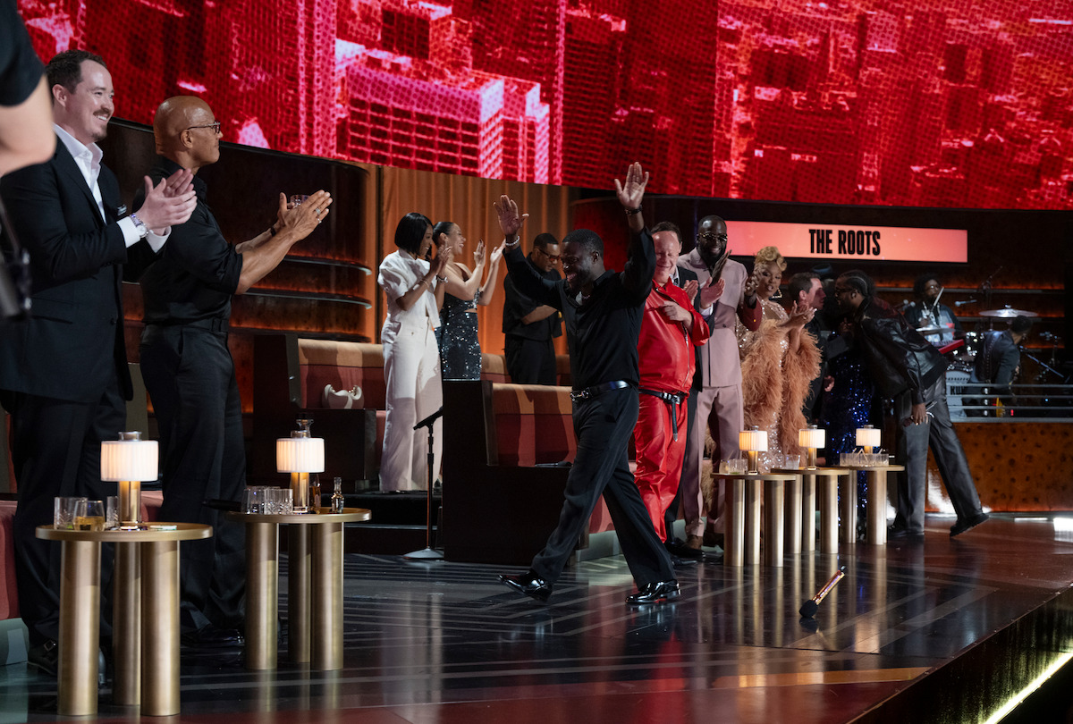 A group of people dressed formally on a stage with tables and small lamps, clapping and cheering in front of a large screen displaying "The Roots," in an elegant event or award show setting.