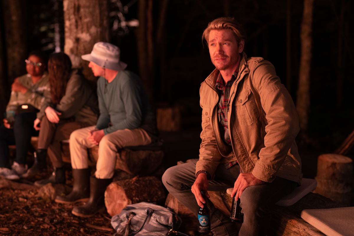 In an image from ‘Sullivan’s Crossing,’ Chad Michael Murray holds a bear while wearing a jacket as he sits on a bench in front of the glow of a fire.