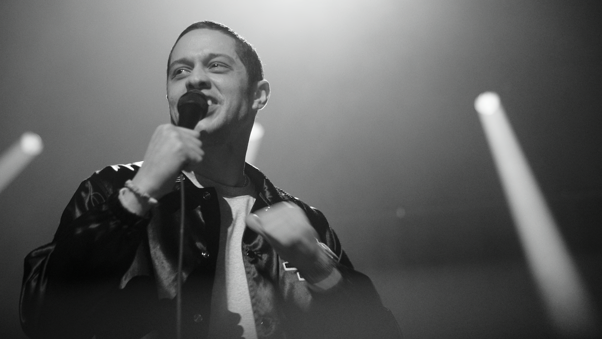 A man holding a microphone performs onstage in a dark, smoky environment with dramatic lighting and beams, appearing to be engaged with the audience.
