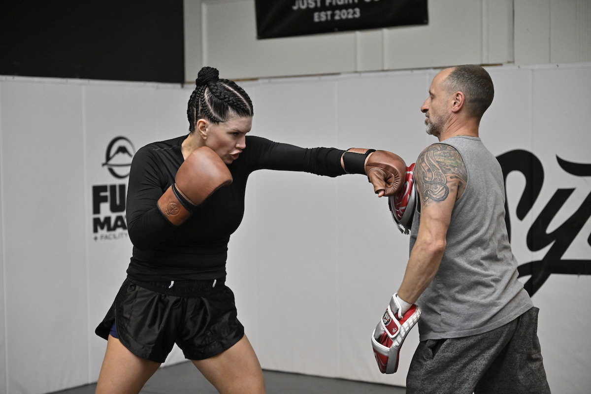 A female boxer with braided hair practices punching with a male trainer holding pads in an indoor gym, surrounded by white padded walls and gym logos, both focused and in athletic gear.