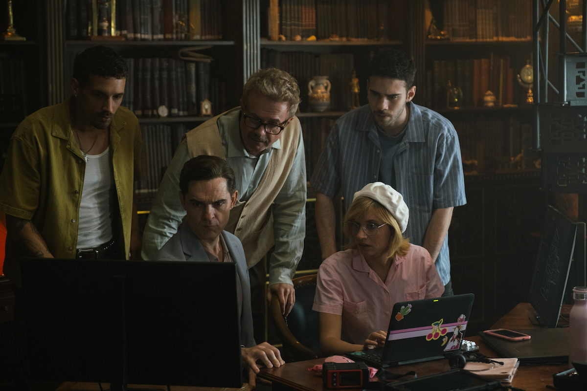 Five people in a library or study room, gathered around two computer monitors with bookshelves in the background, appearing focused and engaged in collaborative work or problem-solving.