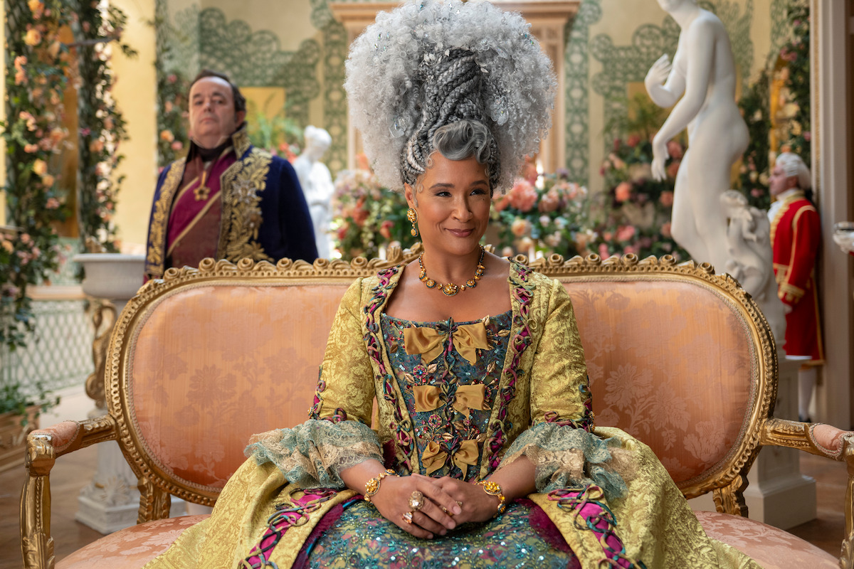 Woman in elaborate historical costume sits on a luxurious sofa in a richly decorated room with flowers, statues, and another person in period attire behind her.