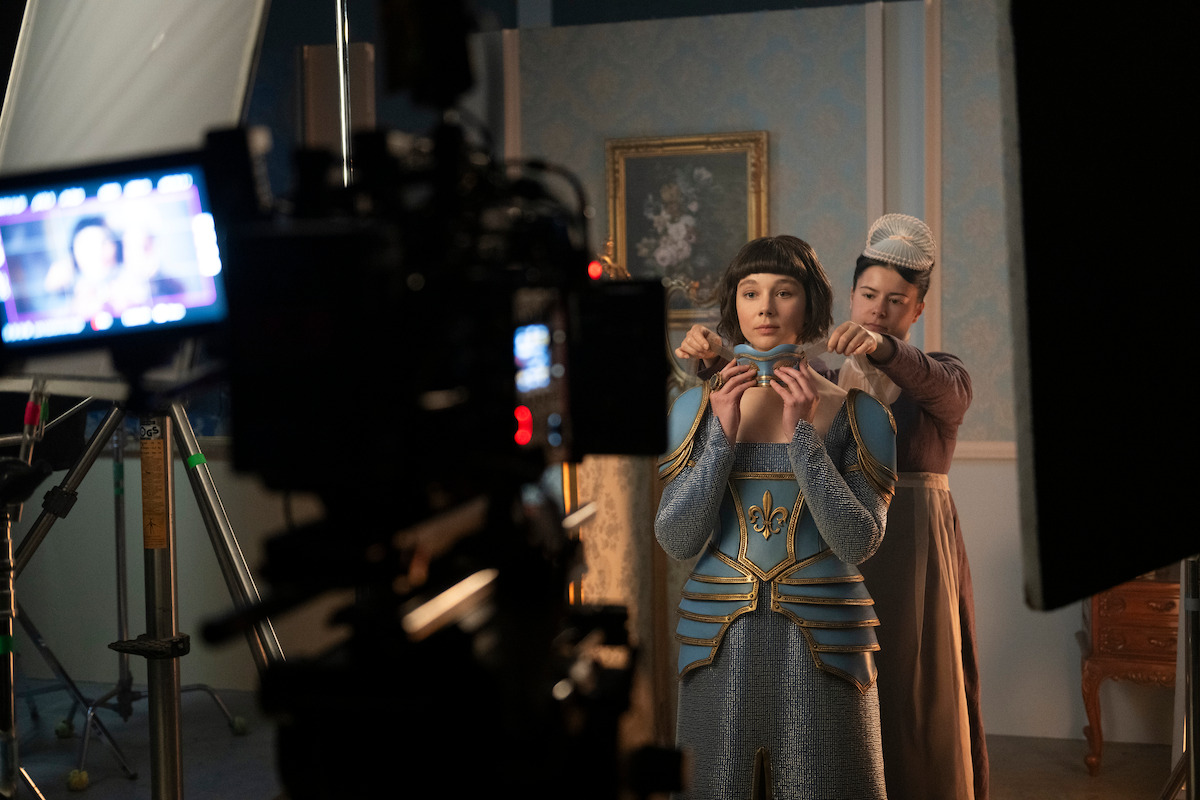 A film set with a woman being assisted with her costume by another woman, surrounded by studio lights and camera equipment in an ornate, vintage room.