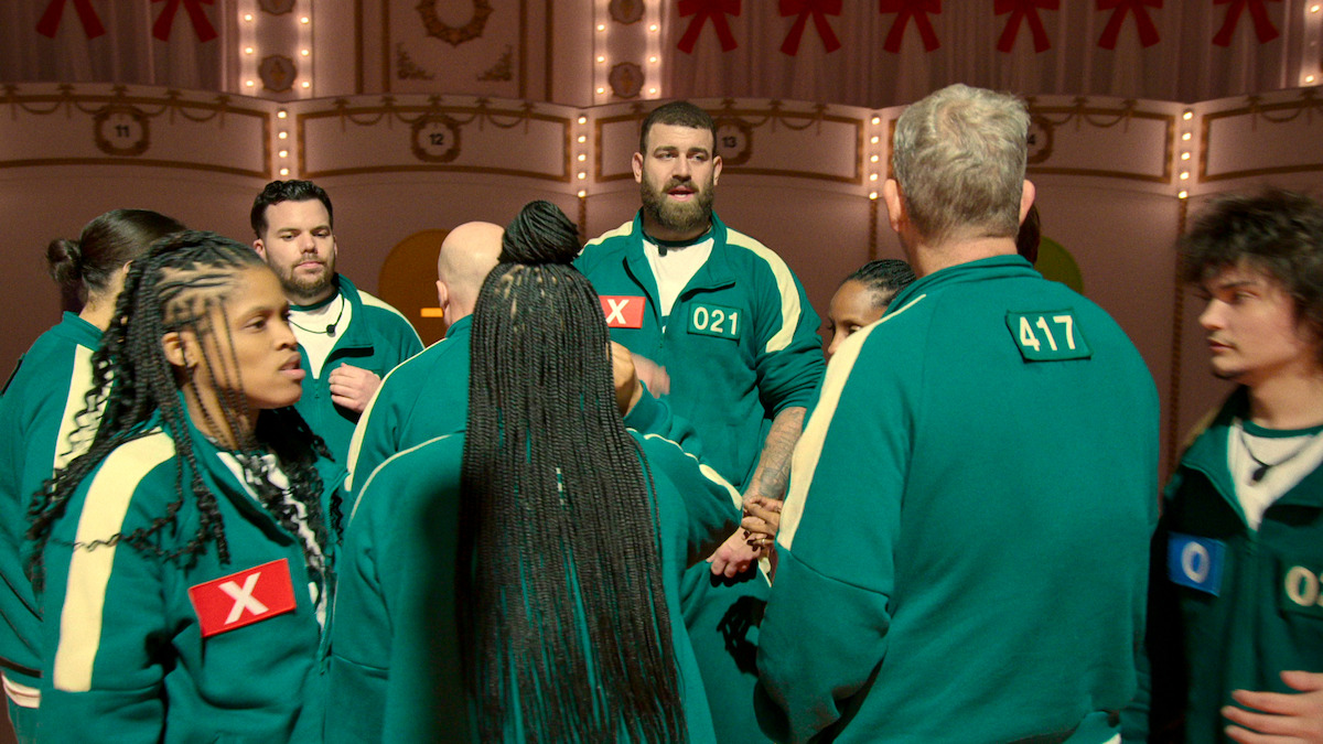 A group of contestants in green tracksuits stand in a lit game arena, some marked with Xs, gathered in a tense huddle, suggesting anticipation and competition in a high-stakes environment.