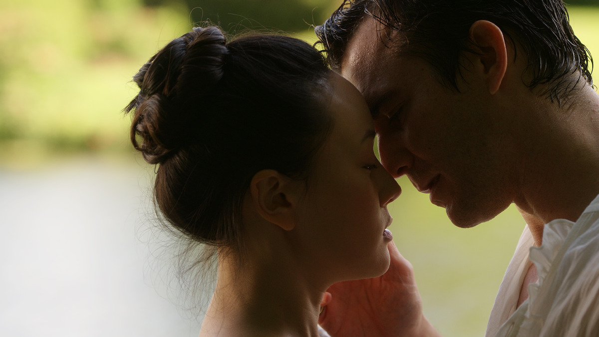 A couple stands close together by a serene lake, gently touching foreheads in an intimate moment, with soft natural light and greenery in the background.