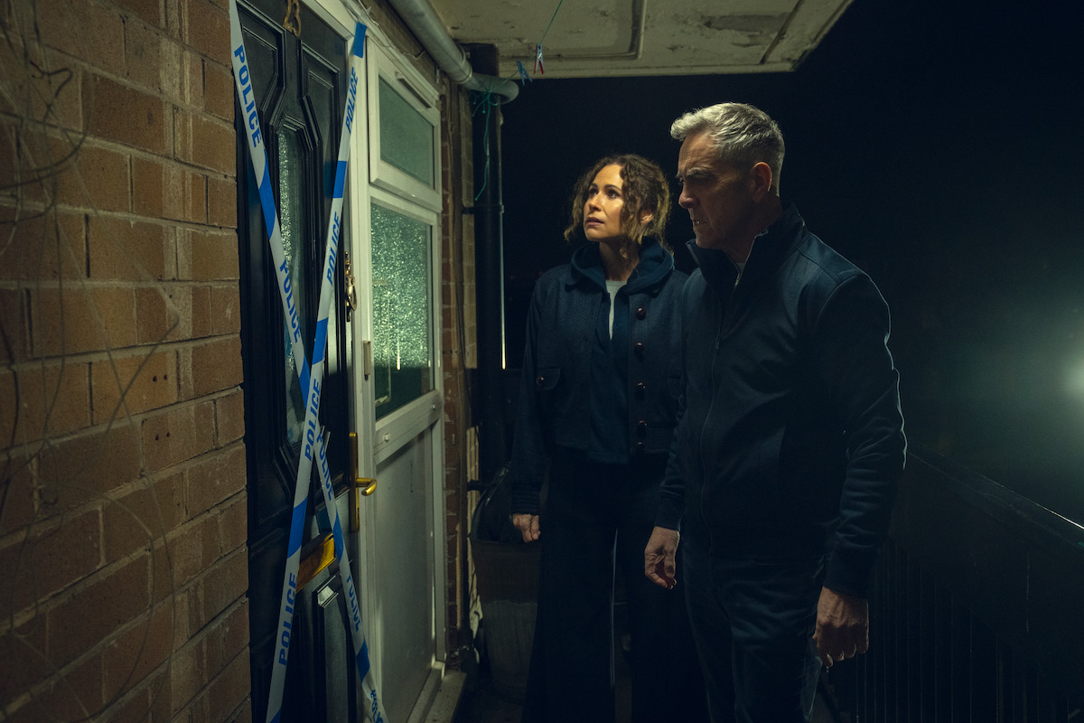Two detectives stand outside a taped-off crime scene door at night, looking serious and focused, illuminated by harsh lighting on a brick-walled balcony area.