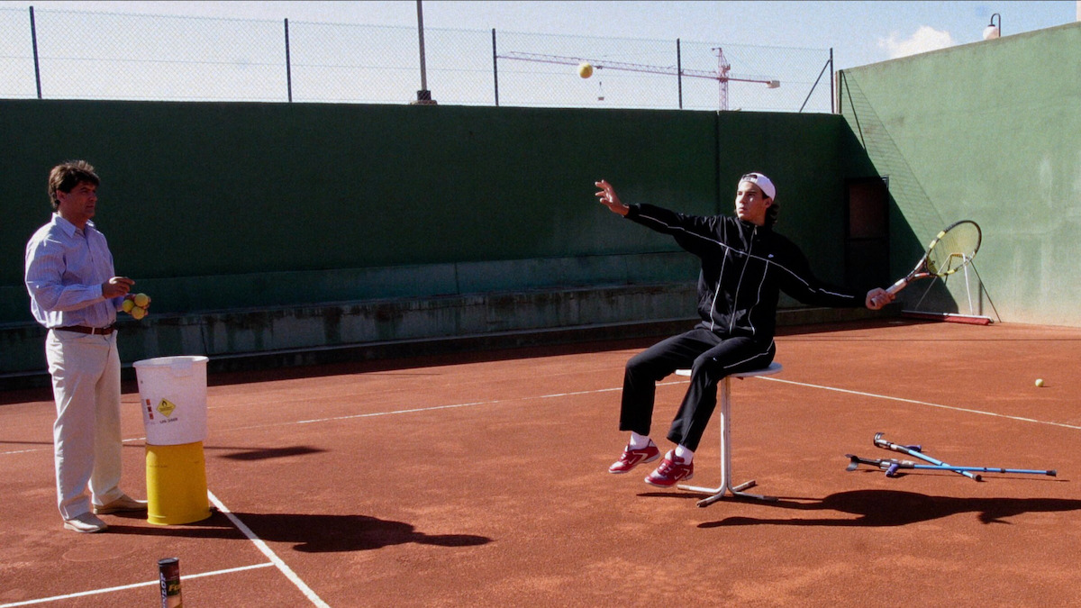 A tennis player practices serving while seated on a chair on a clay court, with a coach observing and holding tennis balls. Extra racquets and a can lie nearby, and the court is bordered by green walls and a wire fence.