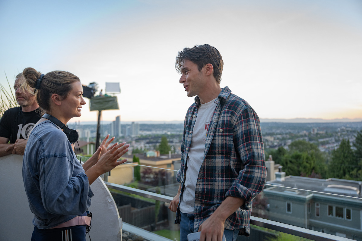 Three people talking on a rooftop balcony at sunset, overlooking a cityscape. One woman gestures as she speaks to a man in a plaid shirt, while another person stands in the background holding a reflector. Casual, outdoor urban setting.