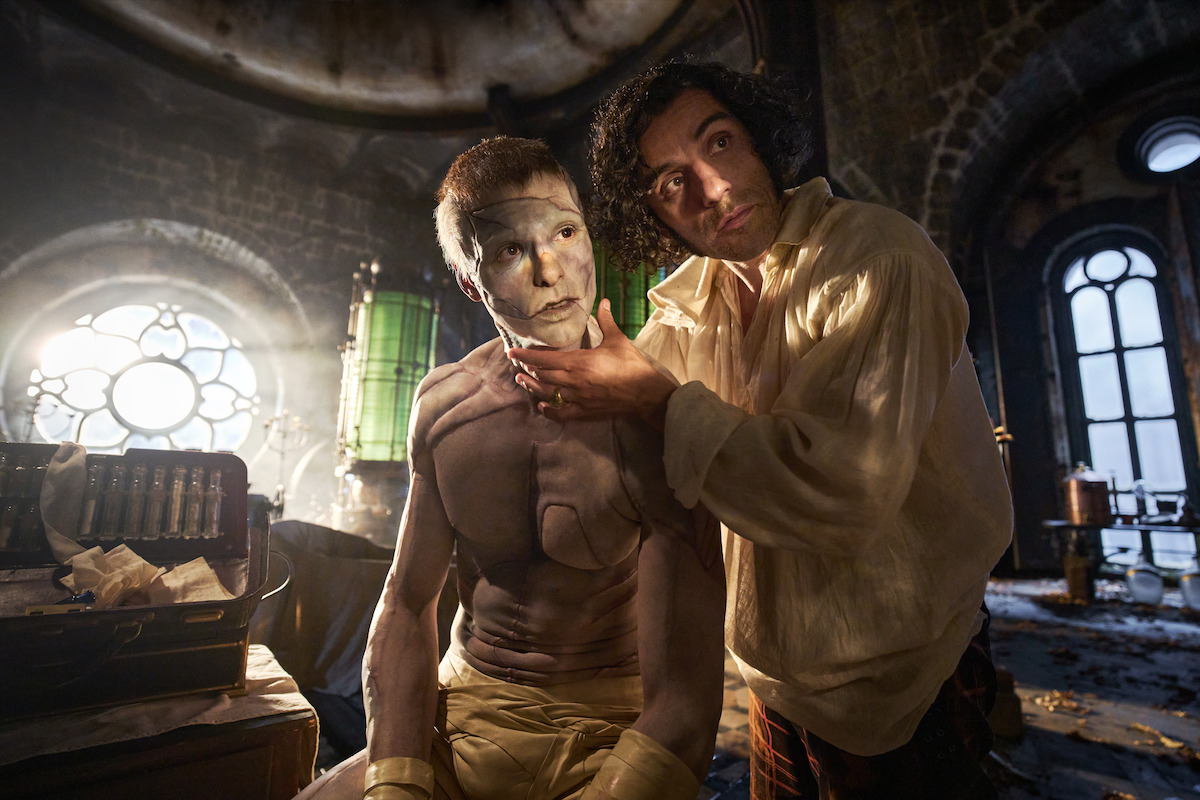 A man applies white makeup to a shirtless figure in a dim, gothic laboratory bathed in dramatic light, evoking a mysterious, tense, and atmospheric mood with vintage scientific equipment in the background.