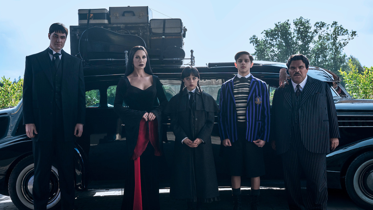 Joonas Suotamo as Lurch, Catherine Zeta-Jones as Morticia Addams, Jenna Ortega as Wednesday, Isaac Ordonez aș Pugsley Addams, Thing, and Luis Guzmán as Gomez Addams stand in front of their car in Season 2 of ‘Wednesday’