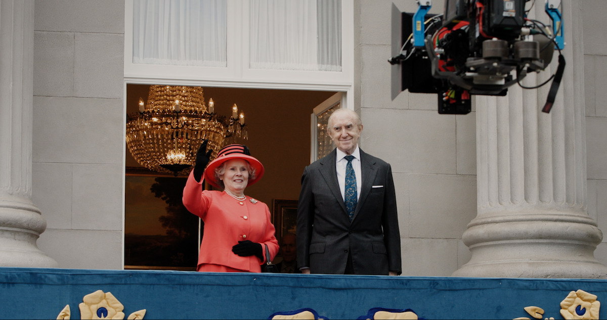 Behind the scenes shot of Imelda Staunton as Queen Elizabeth II and Jonathan Pryce as Prince Phillip waving on a palace balcony in Season 6 Part 2 of 'The Crown'