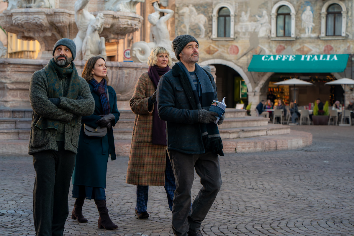 Four adults in winter clothing walk together in a European plaza near a fountain, with a café and people dining outside in the background, suggesting a casual sightseeing outing in an urban historic setting.