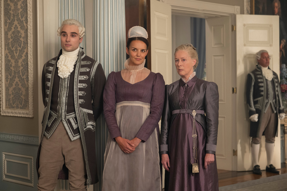 Three people in period costumes stand indoors in an ornate historical room, with detailed wallpaper, woodwork, and another person in the background near an open door.