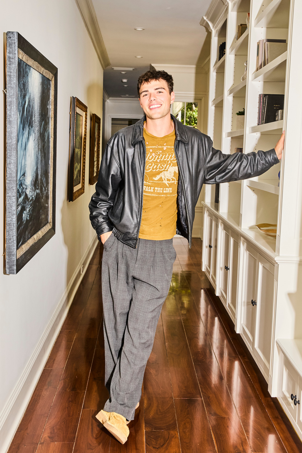 A smiling young man in a leather jacket, casual trousers, and moccasins stands relaxed in a bright hallway with hardwood floors, framed art on the wall, and built-in bookshelves.