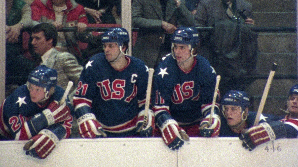 US hockey players in blue uniforms with "USA" on the front sit and stand on the team bench during a game, holding hockey sticks; audience members are visible in the background, creating an intense sports atmosphere.
