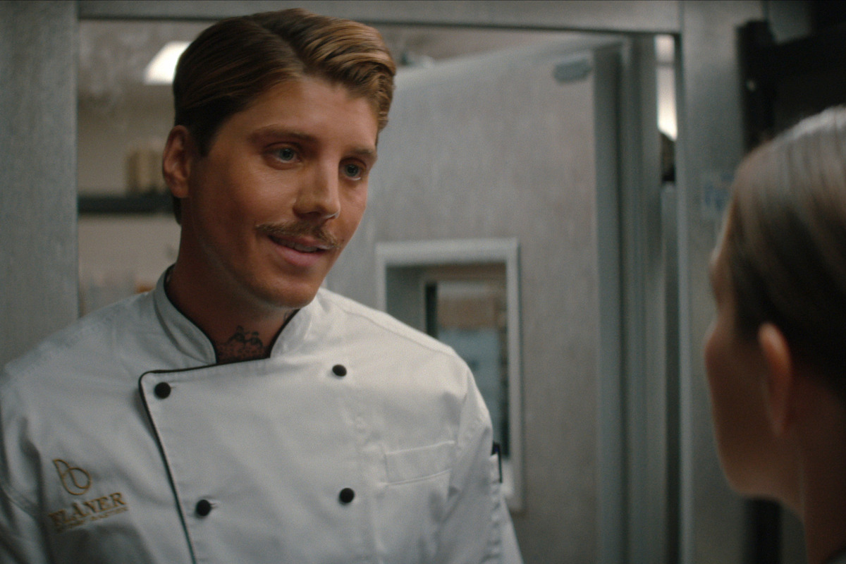 Male chef in a white uniform with embroidered name speaks to a woman in a commercial kitchen setting, stainless steel appliances and open door visible in background.