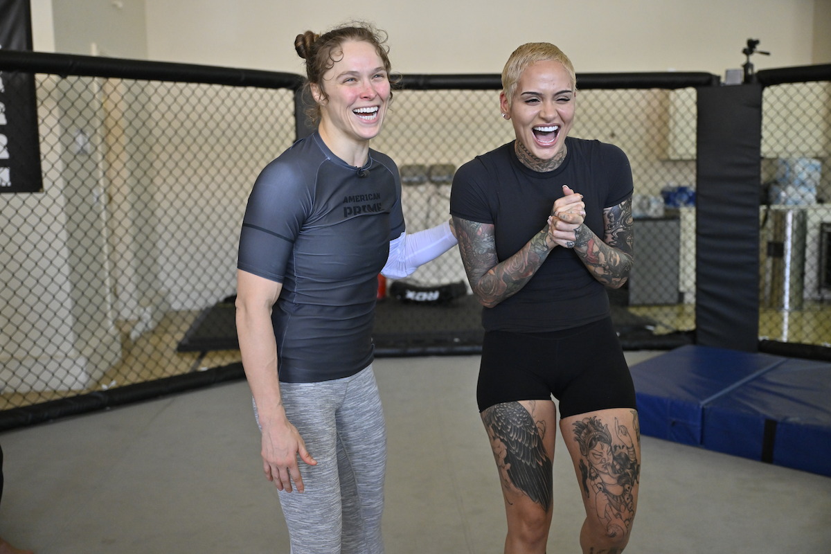 Ronda Rousey and Kehlani laughing together in an MMA training gym, standing on mats near a fenced cage, wearing athletic workout clothing with one sporting visible tattoos.